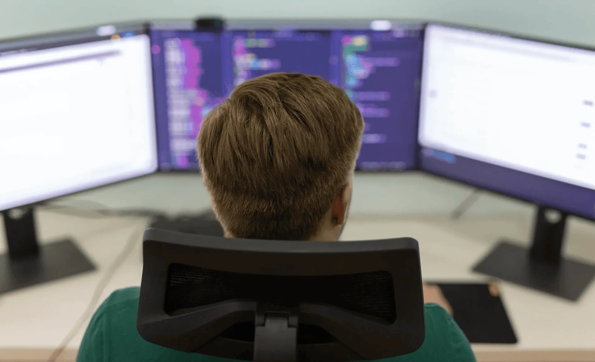 A photo of a person sitting behind a computer having three monitors open, coding